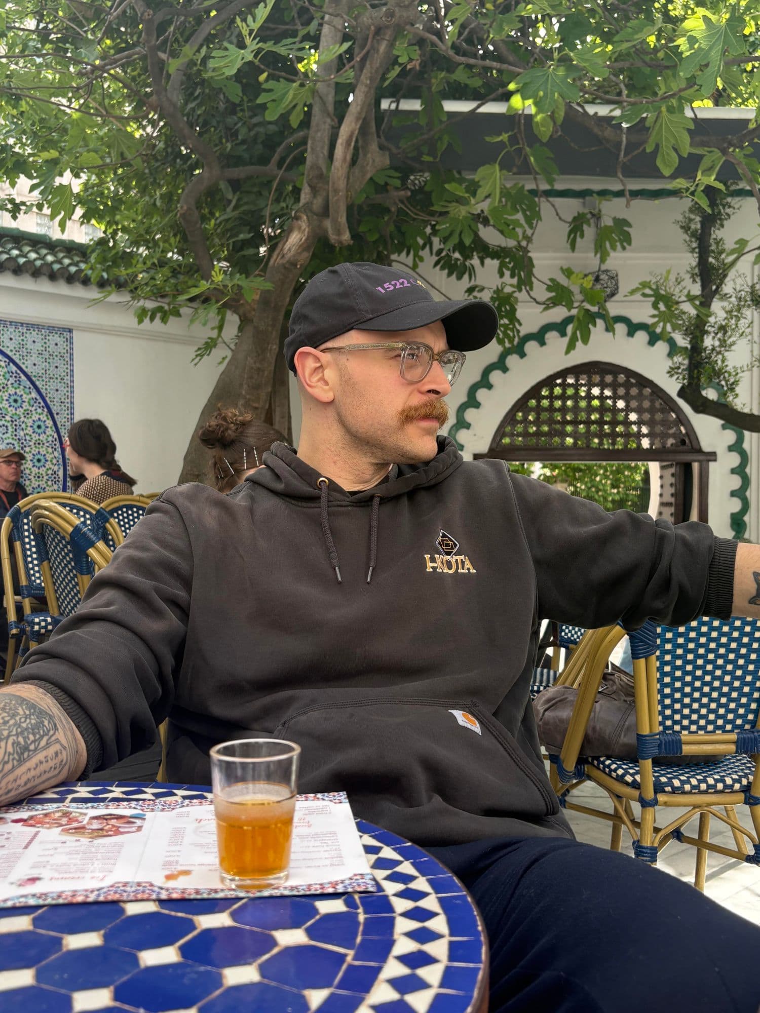 Filip in a cap and hoodie at a mosaic café table with a glass of tea, in a tiled courtyard under a fig tree.