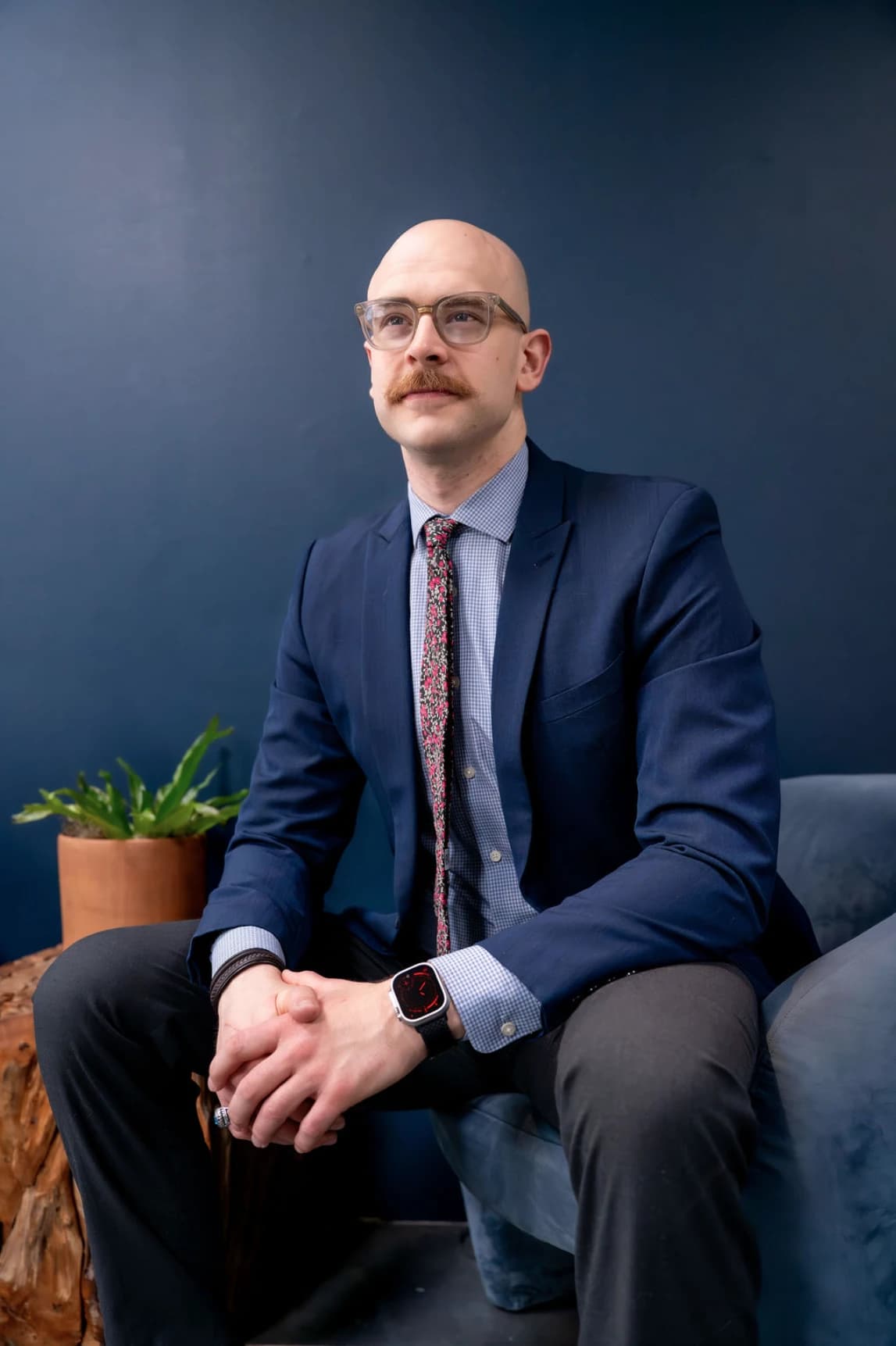 Filip in a navy suit, seated against a dark blue wall and looking up and to the side.